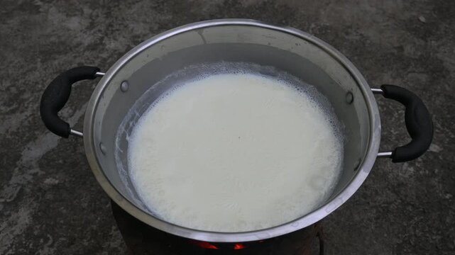 Fresh milk is boiling in a large stainless steel vessel. It has been placed over a traditional fire burning outdoors. As the milk heats up, foam begins to form along its edges. 