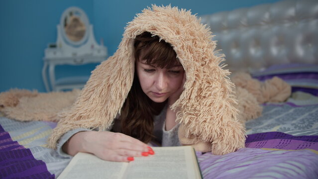 brunette woman lies on the bed and reads a book, tited and covered with a blanket, have fun and enjoy