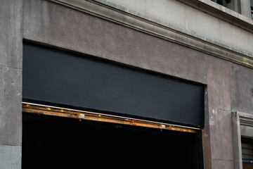 Building facade showing a worn concrete wall texture with a prominent dark storefront sign blank for text, revealing a partially open old metal security gate and a dark void inside © Volodymyr_sh