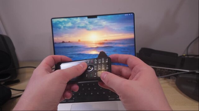 Hands using old keypad mobile phone in front of modern laptop computer screen, illustrating contrast between early mobile communication devices, contemporary digital technology in everyday workspace