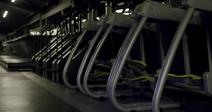 Modern fitness club interior with rows of elliptical cross trainers and treadmills in a dark, empty gym, highlighting the sleek design of the equipment and creating a moody, athletic atmosphere