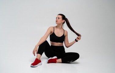 Fototapeta premium Fitness woman sitting on white background, wearing black sportswear and red sneakers, pulling her long ponytail, demonstrating a healthy and active lifestyle