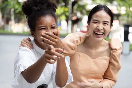 Two young women, Asian and African friends having a good time, talking and laughing together, pointing at you