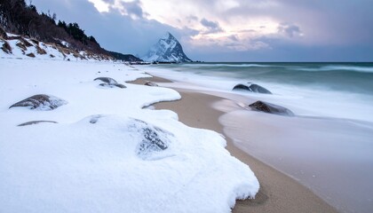 Fototapeta premium Snow-covered beach with ocean and mountain under cloudy sky, winter landscape.
