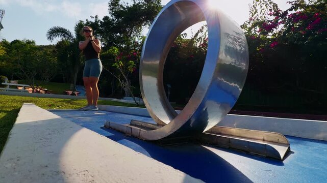 Woman playing mini golf hits ball accurately through circular metal obstacle on outdoor tropical course in bright sunlight