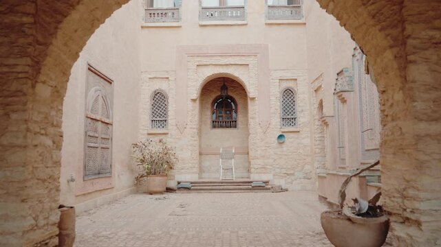 Stone archway entrance to a Moroccan courtyard with traditional architecture and decor. The courtyard features plants, pots, and a chair, creating a peaceful ambiance.