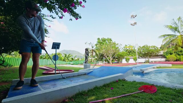 Man Hits Golf Ball Over Ramp Above Water Obstacle on Mini Golf Course in Tropical Park, Wide Angle View of Player Attempting Difficult Shot With Putter Near Blue Ramp and Pond, Leisure Sport Challenge