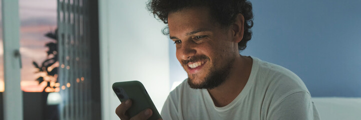 Fototapeta premium Guy is sitting on the bed in the bedroom, watching video, movie on mobile phone. Panoramic