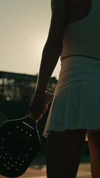 A vertical, close-up shot from behind a female athlete standing on a pickleball court during golden hour. The woman is wearing a white athletic skirt and tank top, holding a black pickleball paddle.