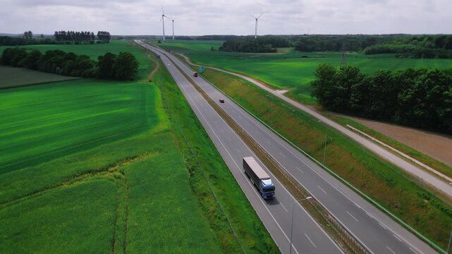 Aerial shot of long-haul truck traveling along a vast highway, passing a sprawling wind farm with towering turbines spinning gracefully, clean energy powering the future of transport. 4k footage
