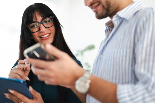 A woman wearing glasses and a man in a striped shirt collaborate while looking at a mobile device and tablet in an office setting.