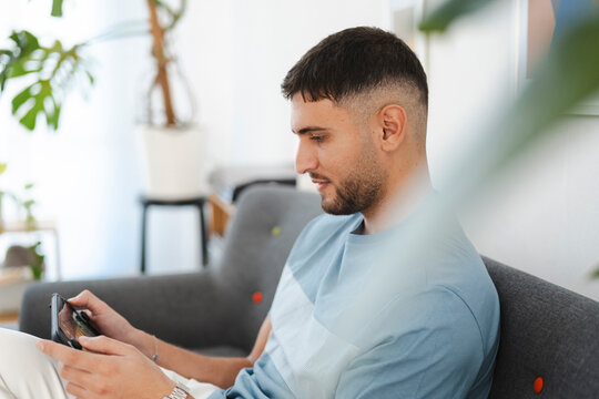 A young man sits on a sofa, engrossed in playing a video game on a handheld console. He is focused on the screen, his thumbs actively controlling the game.