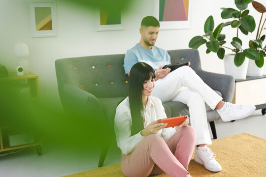 A young couple enjoys playing video games together in a modern living room. The woman sits on the floor with an orange handheld console, while the man relaxes on the sofa with a black one.