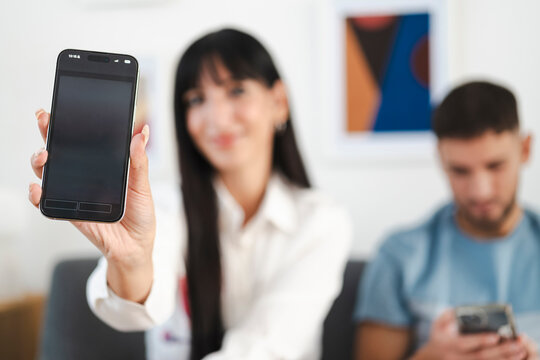 A woman holds a smartphone displaying an event app, while a man in the background also looks at his phone.