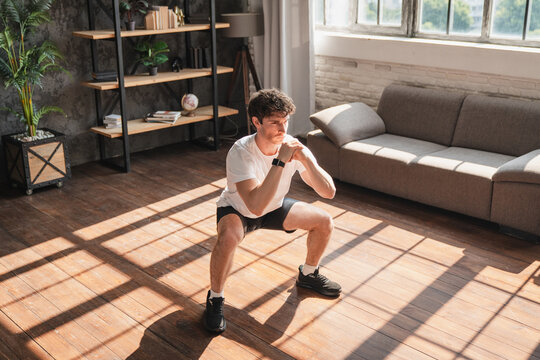 Young Caucasian man doing bodyweight squats at home, training legs to strengthen quadriceps, hamstrings, and glutes. Home fitness workout for lower body strength, balance, and wellness.