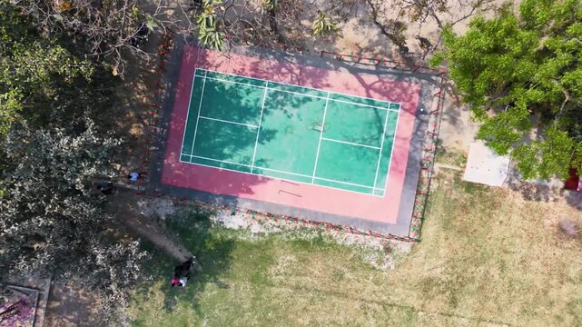 Aerial view of empty outdoor green tennis court with red border in sunny park surrounded by trees and fence