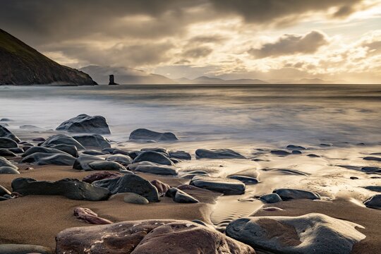 Sandy beach beach with stones on North Atlantic with rocky outcrop and cloudy sky, Dingle Peninsula, Kerry, Ireland