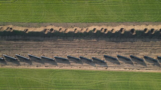 Top-down aerial view of the Balkan Stream gas pipeline construction in Bulgaria. Steel pipes laid in a row between green agricultural fields and plowed soil