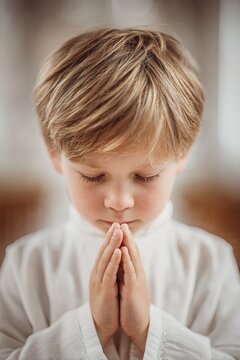 A child is praying with hands clasped together during first communion. The setting is a church with light filtering in and family members gathered nearby