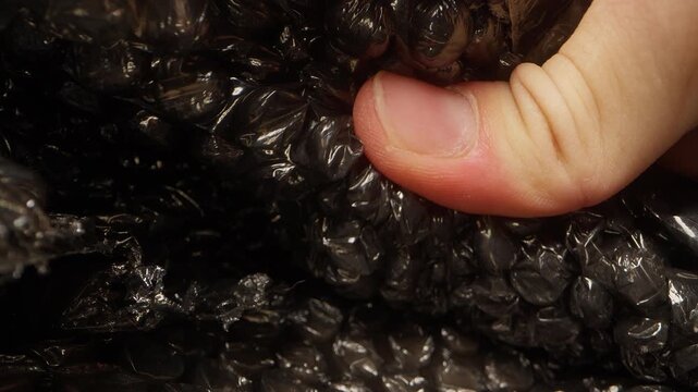 Macro dolly slider shot of a finger squeezing and popping bubbles on black plastic bubble wrap packaging. Detailed cinematic close up of protective packing material compressing under human touch with 