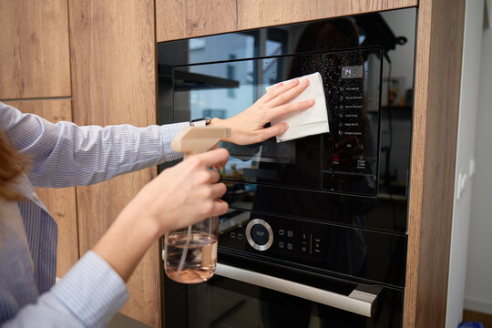 Woman wipes glass door of built in microwave oven while holding spray bottle in kitchen cabinet