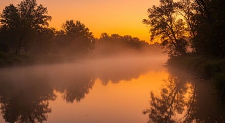 Fototapeta premium Tranquil river surface covered in low-lying mist during a vibrant sunrise reflects surrounding dark foliage.