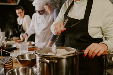 Middle aged Caucasian woman straining cooked rice over pot in foreground, multiethnic group of adults and chef preparing food together in background during cooking class