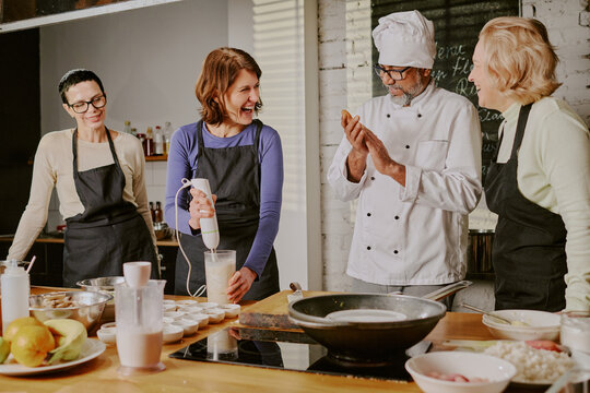 Group of middle aged Caucasian women and chef preparing food together in cooking class, women smiling and interacting while chef demonstrating technique at kitchen counter