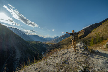Back view of happy hiker with backpack raising arms in success standing on mountain edge overlooking scenic alpine valley with river during autumn trekking trip for freedom concept.
