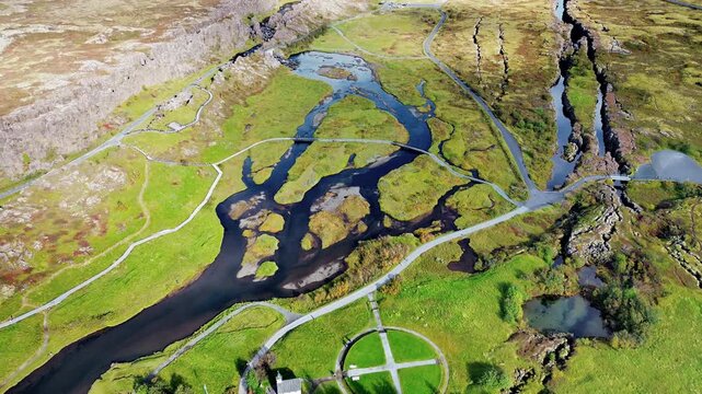 Aerial drone view of the Pingvellir National Park rift valley where tectonic plates separate. Rivers and wetlands weave through green plains surrounded by dramatic rock cliffs and walking paths