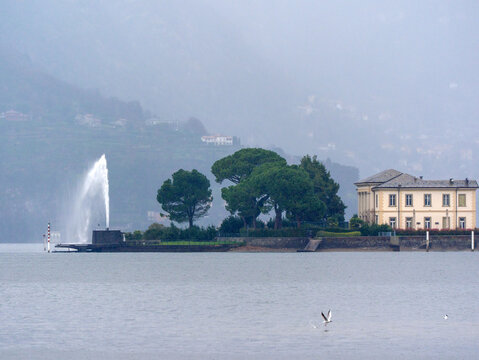 View of Lake Como with Villa Olmo and a water fountain jet near the shoreline, surrounded by trees and mountains fading into mist, capturing the calm atmosphere of northern Italy.
