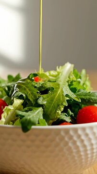 A person pours dressing on a fresh salad with greens and tomatoes in a bowl indoors.