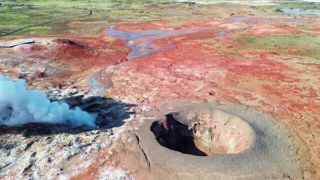Aerial drone view of steaming geothermal vents and bubbling mud pools in a volcanic geothermal field in Iceland. Mineral rich ground in shades of beige, red, and blue surrounds active fumaroles releas