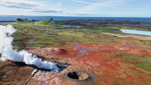 Aerial drone view of steaming geothermal vents and bubbling mud pools in a volcanic geothermal field in Iceland. Mineral rich ground in shades of beige, red, and blue surrounds active fumaroles releas