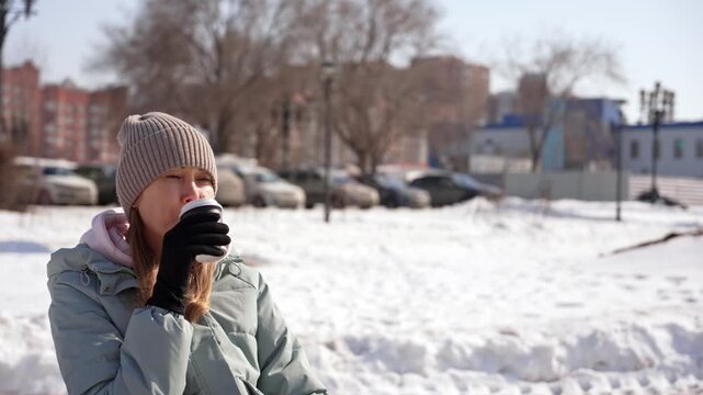 Thoughtful woman wearing a warm jacket, hat, and gloves drinking a hot beverage from a paper cup while relaxing outdoors in a city park on a sunny winter day, enjoying the cold weather