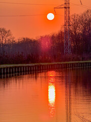 Fototapeta premium Dramatic sunset with electrical transmission tower silhouette reflected in calm water with vibrant orange sky