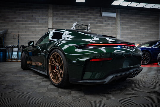 Deep green Porsche 911 GT3 Touring sits in a workshop with cinderblock walls, fluorescent skylights, patterned garage floor, quad exhaust, LED taillight bar, and bronze wheels.