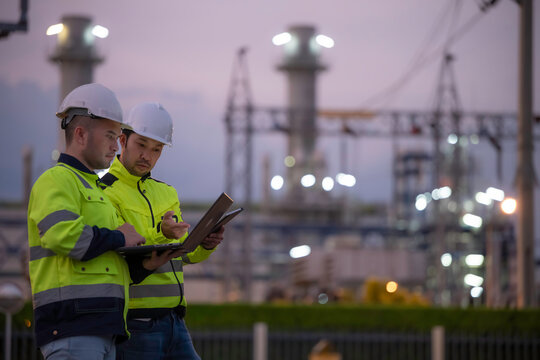 Professional engineers using a digital tablet at a petrochemical refinery or oil and gas power plant during twilight. Concept of energy industry, petroleum infrastructure, and industrial teamwork.