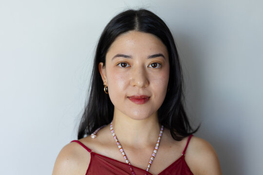 Asian female posing against light gray wall wearing maroon strappy top, beaded necklace, gold hoops