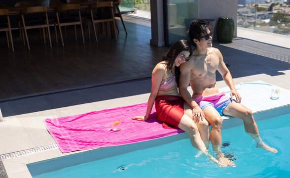 Diverse couple sitting at rooftop pool edge on pink towel wearing sunglasses swim trunks and sarong