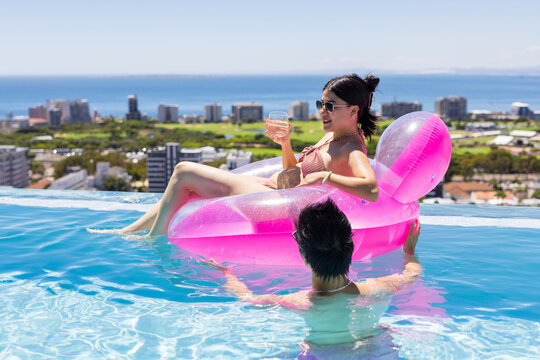 Reclining woman bikini holding stemless glass on pink float at rooftop infinity pool over ocean