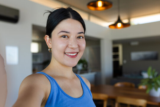 Asian woman taking selfie, smiling in home kitchen in blue tank top with smartphone, potted plant