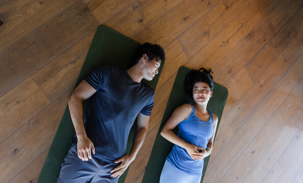 Diverse couple lying on dark green exercise mats on warm wooden floor, woman wearing fitness band