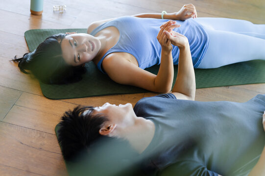 Asian couple lying on green yoga mats on wooden floor holding hands near water bottle