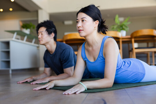 Asian couple practicing prone yoga in living room on yoga mats, woman wearing blue tank top