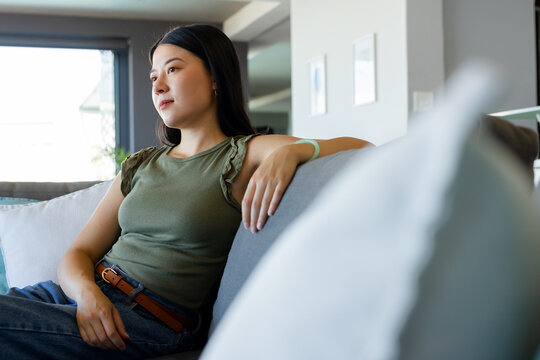 Asian woman sitting on gray sofa in living room wearing olive top, jeans, gazing out window