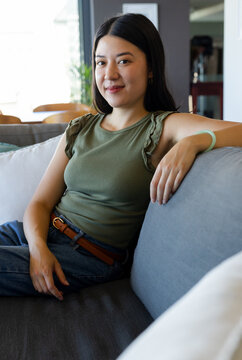 Asian woman sitting on grey sofa at home, wearing olive-green top, hoop earrings visible