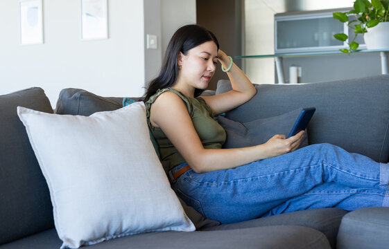Adult Asian woman reclining on gray sofa in living room, wearing olive top, jeans, holding phone