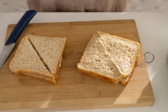 Wooden board holding two wheat sandwiches and blue knife on white counter, light casting shadow