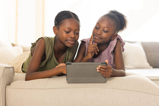 African American sisters, youth, child lying on sofa in living room using tablet in folio case